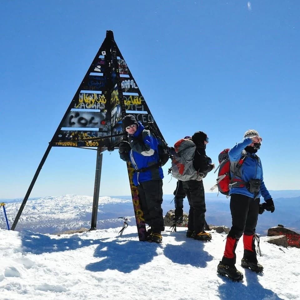 Trekkers in winter gear beside the Toubkal summit marker, snow underfoot