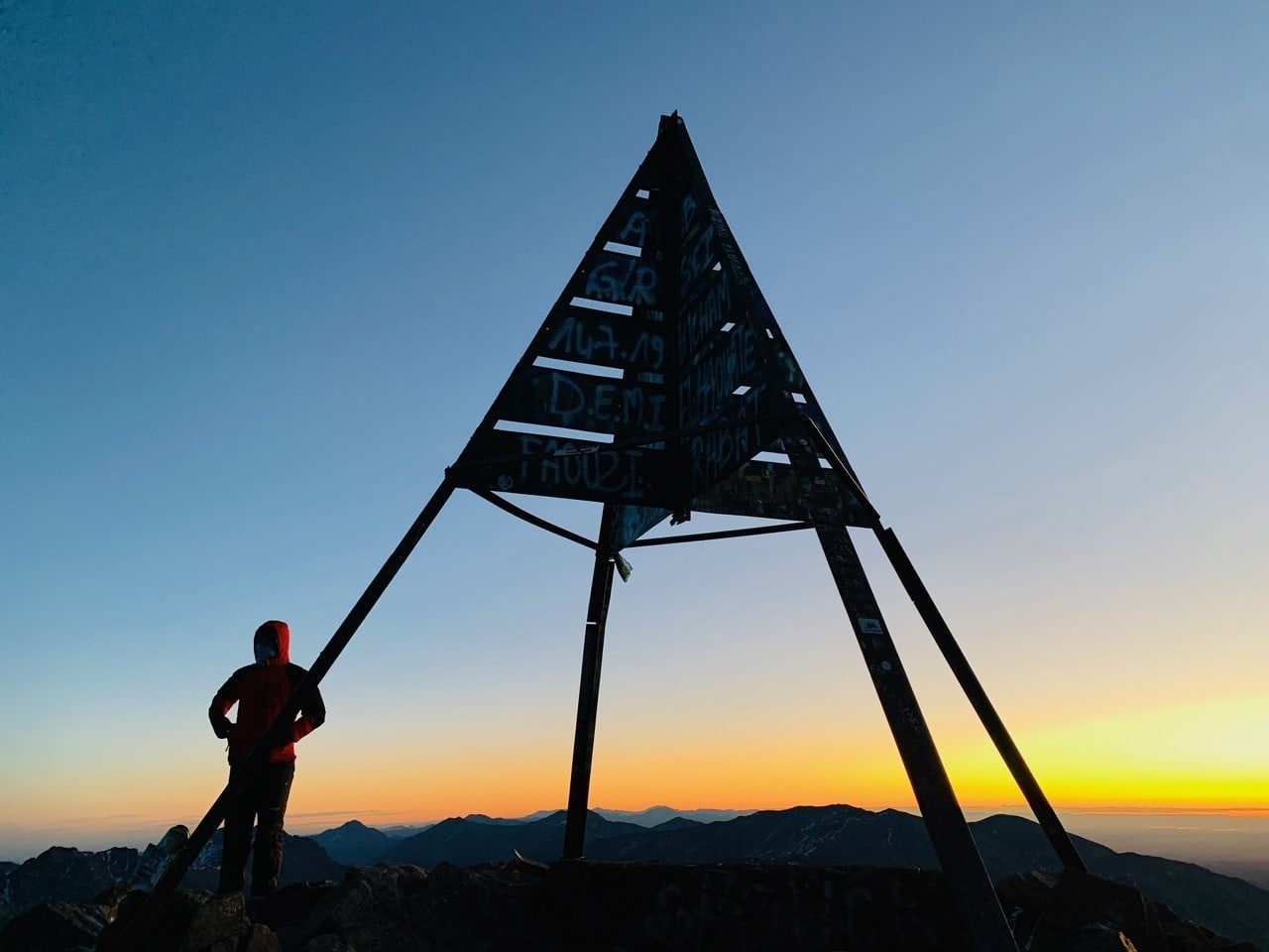 A trekker beneath the summit marker of Mount Toubkal at sunrise, the Atlas Mountains stretching to the horizon